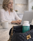 Woman using a laptop with a bottle of 'Vibrant Soothe & Move' supplements in a black handbag.