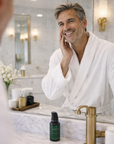 Man in a white robe applying skincare product in front of a mirror with skincare products on the counter.