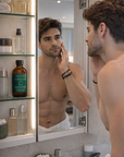 Man applying skincare product in front of a mirror with bathroom products on shelves.