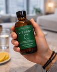Hand holding a bottle of 'Vibrant Skin Vibrant C' dietary supplement in a kitchen setting.