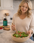 Woman preparing a salad in a kitchen with a bottle of vitamin supplements on the counter.